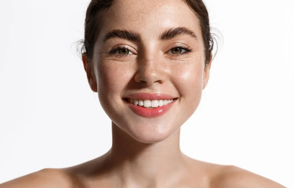Woman with light skin and freckles smiles, with glossy lips, against a white background.