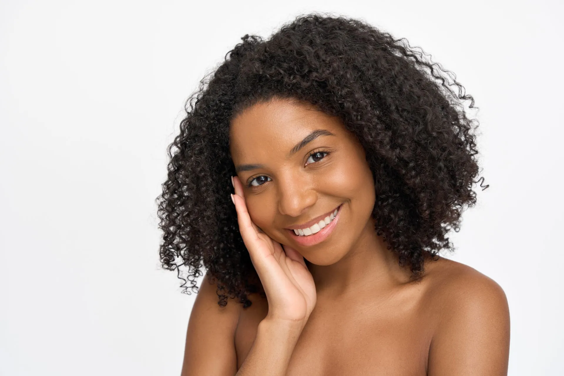 Smiling person with dark curly hair touching their cheek, against a white background.